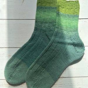 Könnte beinhalten: Ein Paar handgestrickte Socken mit einem Farbverlauf von tiefem Blaugrün an der Spitze und am Fuß bis zu einem helleren Salbeigrün am Schaft. Die Socken weisen eine gerippte Struktur entlang des Fußes und Beins auf und enden in einem limettengrünen Bündchen.