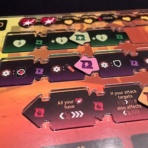 Puede incluir: Primer plano de un juego de mesa con varias piezas. Las piezas son de color púrpura, verde y naranja, con iconos que representan corazones, escudos y rayos. El texto en las piezas incluye "All your have" y "If your attack targets".