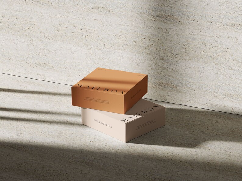 May include: Two square cardboard boxes stacked on top of each other. The top box is a warm tan color with the word MAILBOX printed in black. The bottom box is a light cream color with the word MAILBOX and the text PACKAGING DESIGN printed in black.