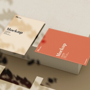 May include: Two blank cards with the word "Mockup" printed on them. The cards are on a light brown surface with a shadow from a plant.