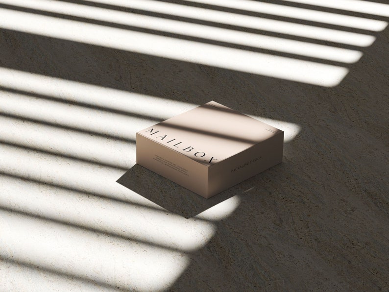 May include: A square, light beige cardboard box with the word MAILBOX printed in black serif font on the top surface. The box features additional small text on the side panels, including the phrase PACKAGING DESIGN, and sits in a light-filled area.