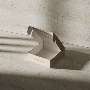 May include: A light beige cardboard shipping box with an open lid. The box features a square base and a hinged top flap. Small, dark, illegible text is printed on the front side of the box, while the interior of the box is a lighter shade.