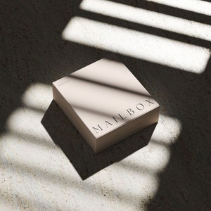 May include: A square, light beige box with the word MAILBOX printed in black capital letters on the top surface. The box has a smooth, matte finish and is positioned at an angle, casting a sharp, dark shadow across the surface beneath it.