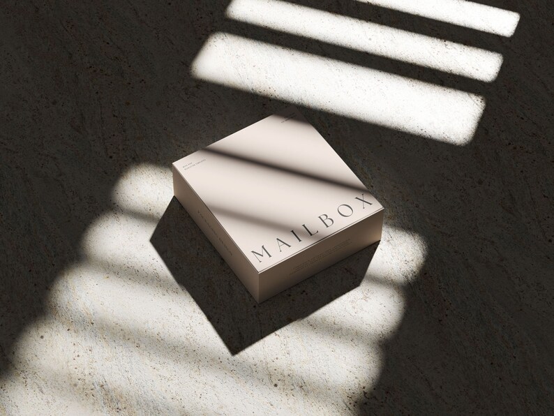 May include: A square, light beige box with the word MAILBOX printed in black capital letters on the top surface. The box has a smooth, matte finish and is positioned at an angle, casting a sharp, dark shadow across the surface beneath it.