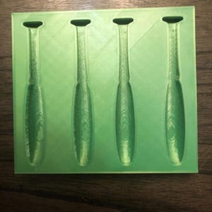 May include: A light green mould featuring four cavities in the shape of baseball bats. The mould is made of a solid material and rests on a wooden surface. The cavities are detailed with the shape of the bats.