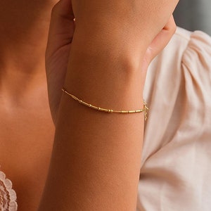 May include: A close-up of a woman's wrist wearing a delicate gold bracelet with small, round beads.