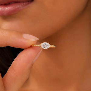 May include: A gold ring featuring a single oval-cut clear gemstone set in a horizontal bezel setting. The band is thin and polished, showcasing a smooth, rounded profile that encircles the finger. The gemstone is faceted and reflects light.