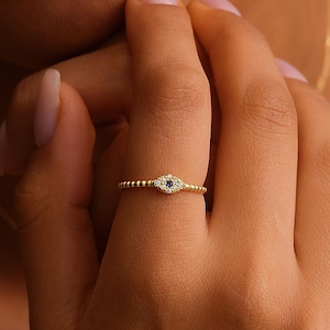 May include: A gold ring with a beaded band and an eye-shaped design. The eye features a small blue stone in the center, surrounded by smaller white stones. The ring is on a finger.