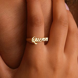 May include: A gold-toned ring featuring the name Quenn written in a cursive script font. The metal band is smooth and polished, with the lettering integrated directly into the top portion of the ring, creating a continuous, singular piece of jewellery.