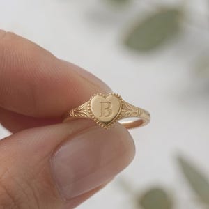 May include: A gold-toned ring featuring a heart-shaped face with a beaded border and an engraved letter B in the center. The band of the ring displays intricate, delicate filigree-style detailing along the sides, extending from the heart.