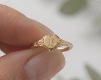 May include: A gold-toned ring featuring a heart-shaped face with a beaded border and an engraved letter B in the center. The band of the ring displays intricate, delicate filigree-style detailing along the sides, extending from the heart.