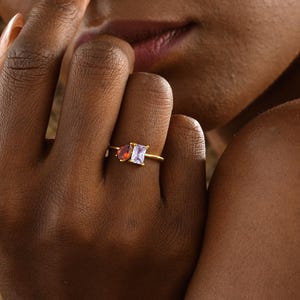 May include: A gold ring with two rectangular gemstones, one purple and one orange. The ring is on a person's finger. The ring's band is thin and gold-coloured. The image is a close-up of the ring.