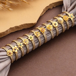 May include: A collection of gold-colored rings, each adorned with a unique floral or gemstone design. The rings are arranged on a gray fabric, with a neutral background. The rings are likely for fashion or jewelry.