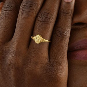 May include: A gold-colored ring with a cross design. The ring features a detailed cross within a circular setting, and is worn on a finger. The ring is made of a shiny, gold-toned metal.