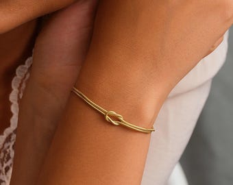 May include: A gold-toned bracelet featuring a delicate snake chain design with a central knot detail. The bracelet is worn around the wrist, showcasing a smooth, metallic finish and a refined, slender profile that wraps securely around the arm.