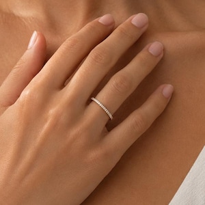 May include: A thin, metallic band ring featuring a continuous row of small, clear, sparkling stones set along the outer circumference. The ring is worn on the ring finger, showcasing a simple, refined design with a polished, reflective finish.