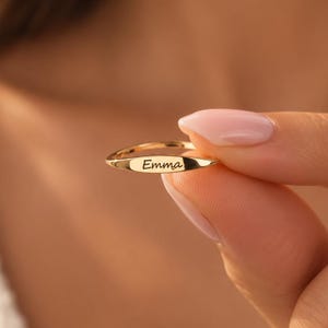 May include: A gold-toned metal ring featuring a thin, polished band and a flat, horizontal bar centerpiece. The bar is engraved with the name Emma in a delicate, cursive script font, centered clearly on the front surface of the ring.