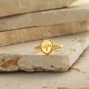 May include: A gold-colored ring with an oval-shaped face featuring an engraved floral design. The ring has a beaded border around the floral design and a detailed band. The ring is displayed on a light-colored stone surface.