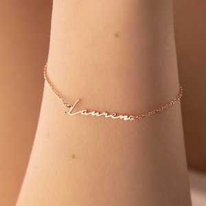 May include: A delicate rose gold bracelet with the name "Lauren" in cursive script. The bracelet features a fine chain and is shown on a person's arm. The name is the focal point of the jewelry.