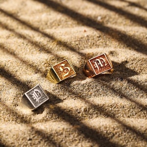 May include: Three square-faced rings with engraved designs rest on sand. One is silver with a stylized letter, another is gold with the number 5, and the third is gold with the letters "JD". The rings are in a sunny setting.