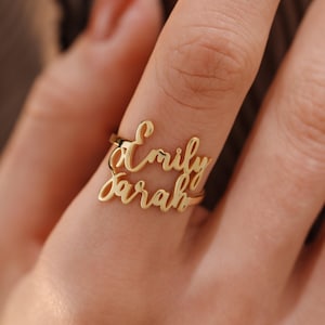 May include: A gold ring featuring the names "Emily" and "Sarah" in a cursive script. The ring is displayed on a finger, highlighting the custom design. The ring is a piece of jewellery.