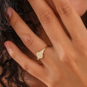 May include: A gold signet ring with a floral design. The ring is worn on a finger, with the design visible. The ring has a detailed floral pattern on a rectangular face. The ring is made of a shiny, gold-colored metal.