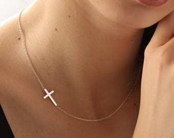 May include: A silver-colored necklace featuring a sideways cross pendant. The delicate chain is draped around the neck, with the cross positioned to the side. The jewelry is simple and elegant.