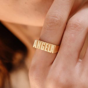 Könnte beinhalten: Ein goldfarbener Ring mit dem Namen "ANGELA" in Blockbuchstaben. Der rechteckige Ring wird an einem Finger getragen. Das Nahaufnahmen-Bild zeigt das Design des Rings und die Haut der Person.