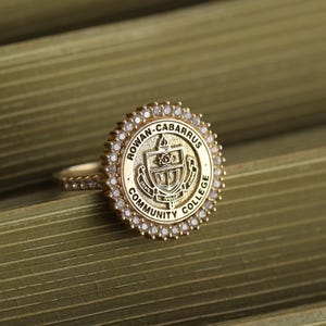 May include: A gold-colored ring featuring the Rowan-Cabarrus Community College emblem. The ring's face is round, surrounded by small, clear stones. The band is also adorned with similar stones, creating a sparkling effect. The ring is set against a green background.