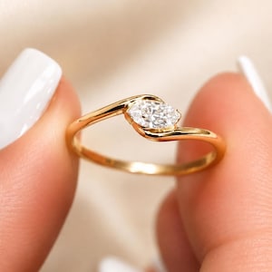 May include: A gold ring featuring a single marquise-cut clear gemstone set in a bypass style band. The polished yellow gold band curves around the elongated stone, creating a sleek and continuous design that highlights the faceted white gem.