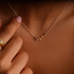 May include: A delicate gold necklace featuring a floral pendant with a small, round, red gemstone at its center. The pendant is attached to a thin gold chain. The necklace is displayed on a person's skin.