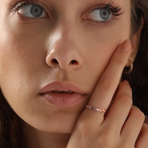 Peut inclure: Une bague en or rose avec un design délicat orné de petites pierres précieuses colorées et d'un détail en forme de cœur. La bague est portée au doigt, mettant en valeur son savoir-faire et son style élégant. C'est un bijou.