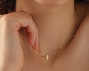 May include: A delicate gold-colored necklace featuring a small cross pendant. The chain is fine and the cross is centered, creating a simple and elegant design. The necklace is worn on a person's neck.