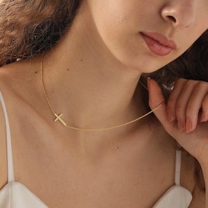 May include: A delicate gold-colored necklace featuring a sideways cross pendant. The cross is positioned to the left of the chain, creating an asymmetrical design. The necklace is worn against a white top.
