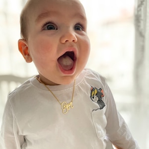 May include: A child wearing a gold-colored necklace with the name "Ryan" in cursive script. The child is wearing a white long-sleeved shirt with a cartoon graphic of Tom and Jerry. The child has an open mouth and is looking upwards.