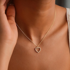 May include: A gold necklace with a heart-shaped pendant. The pendant is adorned with small, sparkling stones. The necklace is worn, highlighting its delicate design. The chain and pendant are gold in colour, creating a subtle sparkle.