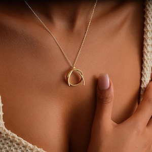 May include: A gold-colored pendant necklace featuring a circular design with intersecting arcs. The necklace hangs on a delicate chain. The jewelry is displayed against a skin-toned background.