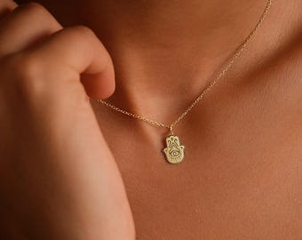 May include: A delicate gold-colored necklace featuring a Hamsa hand pendant. The pendant has an eye symbol in the center. The necklace is displayed on a person's chest.