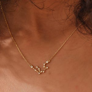 May include: A delicate gold-colored necklace featuring a constellation design. The pendant is adorned with small, sparkling gemstones, creating a celestial aesthetic. The necklace is displayed on a person's skin.