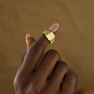 May include: A gold signet ring with a rectangular face is displayed on a finger. The ring has a textured band and a smooth, polished face. The ring is made of a shiny, yellow metal.