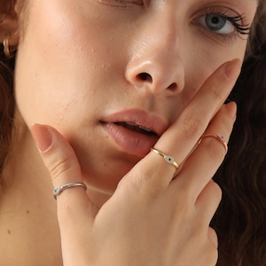 May include: A close-up of a hand displaying three rings. One ring is silver with a small stone, another is gold with a small blue stone, and the third is rose gold with a small stone.