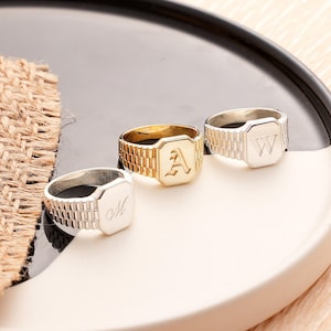 May include: Three signet rings with watch strap-style bands. One gold ring features the letter "A", and two silver rings display the letters "M" and "W". The rings are displayed on a white and black tray.
