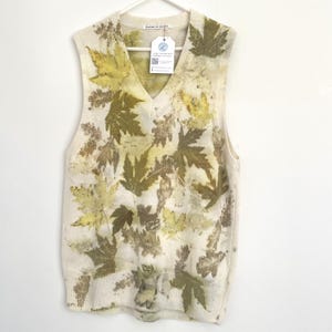 May include: Cream-coloured, sleeveless sweater with a V-neck, decorated with a pattern of green and brown leaves. The leaves are pressed onto the fabric, creating a unique, natural design. The sweater is hanging on a white hanger.