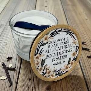 May include: A clear glass jar with a white powder and a blue ribbon inside. The lid, made of bamboo, features the text "GRADWOHL ROAD FARM ALL-NATURAL BODY DUSTING POWDER VANILLA" surrounded by floral illustrations. Vanilla beans are scattered around.