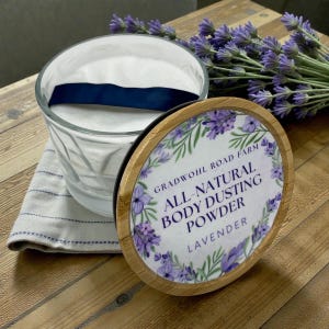 May include: A glass container with a white powder and a blue ribbon, next to a lid with the text "Gradwohl Road Farm All-Natural Body Dusting Powder Lavender". The lid has a bamboo rim and a floral design. A bundle of lavender is in the background.
