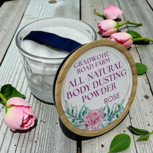May include: A clear glass jar filled with white powder and a navy blue ribbon. The lid, made of bamboo, displays the text "Gradwohl Road Farm All-Natural Body Dusting Powder Rose" surrounded by floral illustrations. Pink roses are scattered around the jar.