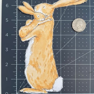 May include: A brown and white rabbit holding a smaller brown and white rabbit in its arms. The rabbits are both looking at the viewer. The larger rabbit has a white tail.