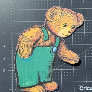 Pu&ograve; includere: Un orsacchiotto di peluche marrone che indossa una salopette blu con un bavaglino verde e un bottone blu. L'orso sta guardando in basso e ha un'espressione amichevole.