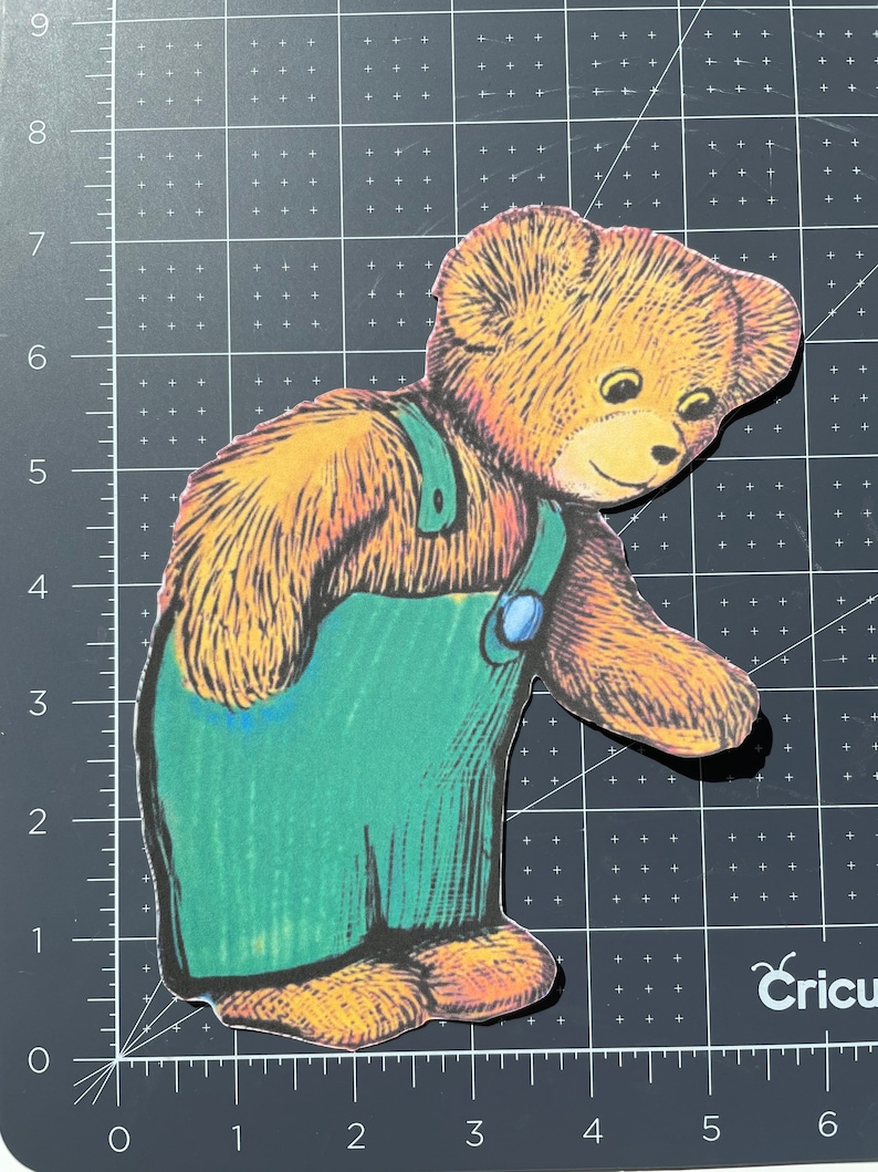 Pu&ograve; includere: Un orsacchiotto di peluche marrone che indossa una salopette blu con un bavaglino verde e un bottone blu. L'orso sta guardando in basso e ha un'espressione amichevole.