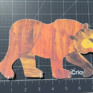 Pu&ograve; includere: Un ritaglio di orso marrone con naso e bocca gialli, che cammina verso destra. L'orso &egrave; fatto di carta con un motivo a venature di legno.